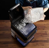 A person in a denim shirt pours a clear container of ice cubes into the open top of a black Waring Ice Crusher, which sits on a wooden surface with some ice already inside the machine.