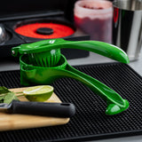 A bright green citrus hand squeezer made of coated zinc alloy sits open on a black bar mat next to a halved lime, mint leaves, and a knife on a wooden cutting board. In the background are red sugar, cherries, and a metal cup.
