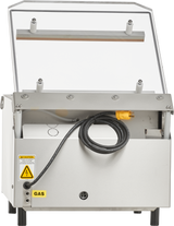 A stainless steel single chamber Vacuum Packaging Machine with a transparent hinged lid open upright. A thick black power cord with a yellow plug is coiled at the front, and gas flush and electric shock warning labels are visible on the lower left side.