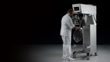 A man dressed in all white operates a heavy duty planetary mixer with a metal whisk attachment, set against a dark background. The stainless steel mixer features a digital control panel as he stands facing it, adjusting the whisk.