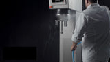 A person in a white chef’s coat sprays water onto a large industrial planetary mixer with a hose. The metallic machine, equipped with control buttons on top, gleams with water droplets contrasting against the dark background. The person’s face is not visible.