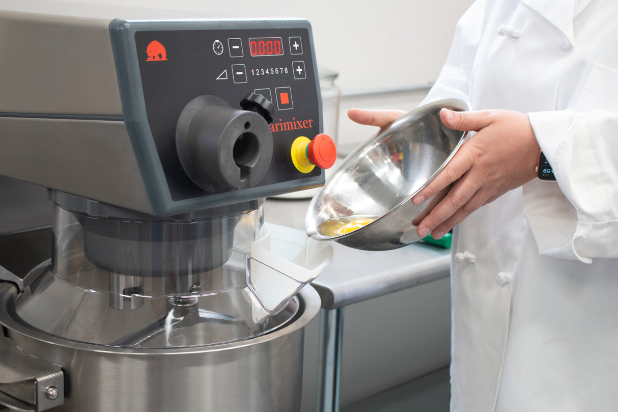 A person in a white chef’s coat pours a yellow liquid from a metal bowl into a large stainless steel food mixer with 20 qt. capacity, featuring control buttons, a red emergency stop button, and digital display in a slightly blurred kitchen background.