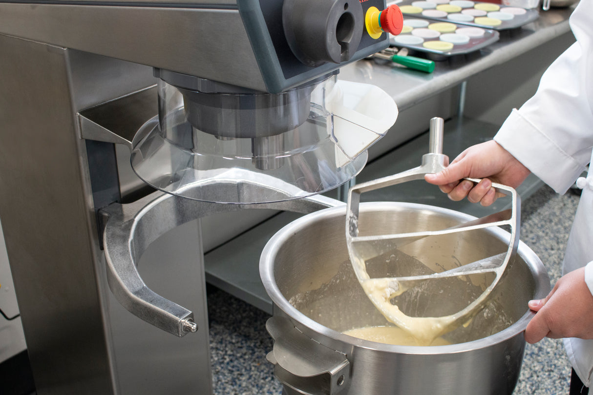 A person in a white coat holds the paddle attachment of a 20 qt. capacity food mixer above a stainless steel bowl filled with yellow batter. Other mixer attachments and trays with baked goods are visible on the counter in the background.