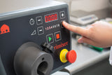A close-up of a person pressing a plus-shaped button on a gray Grimixer food mixer. The digital display shows 0000. The control panel has various buttons, symbols, and a large red emergency stop button. A scale is visible behind the stainless steel unit.