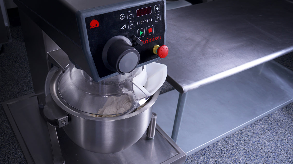 A large Varimixer KODIAK 20 qt. food mixer with a digital control panel and emergency stop button stands on a speckled floor next to a stainless steel worktable. The mixing bowl contains flour, partially covered by a plastic guard.