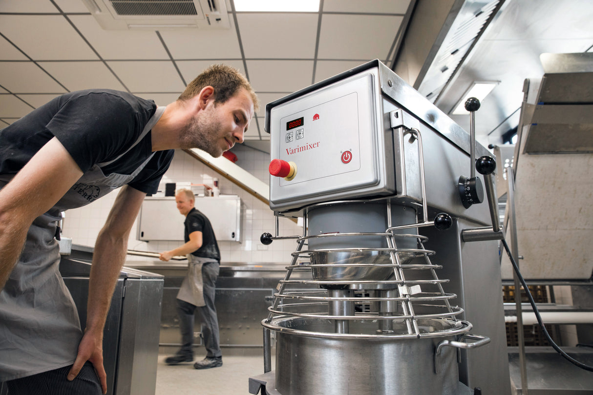 A man in a black shirt and apron leans closely to inspect a large Varimixer V30 planetary mixer in a commercial kitchen. Another person, also in work attire, moves in the background near stainless steel counters.