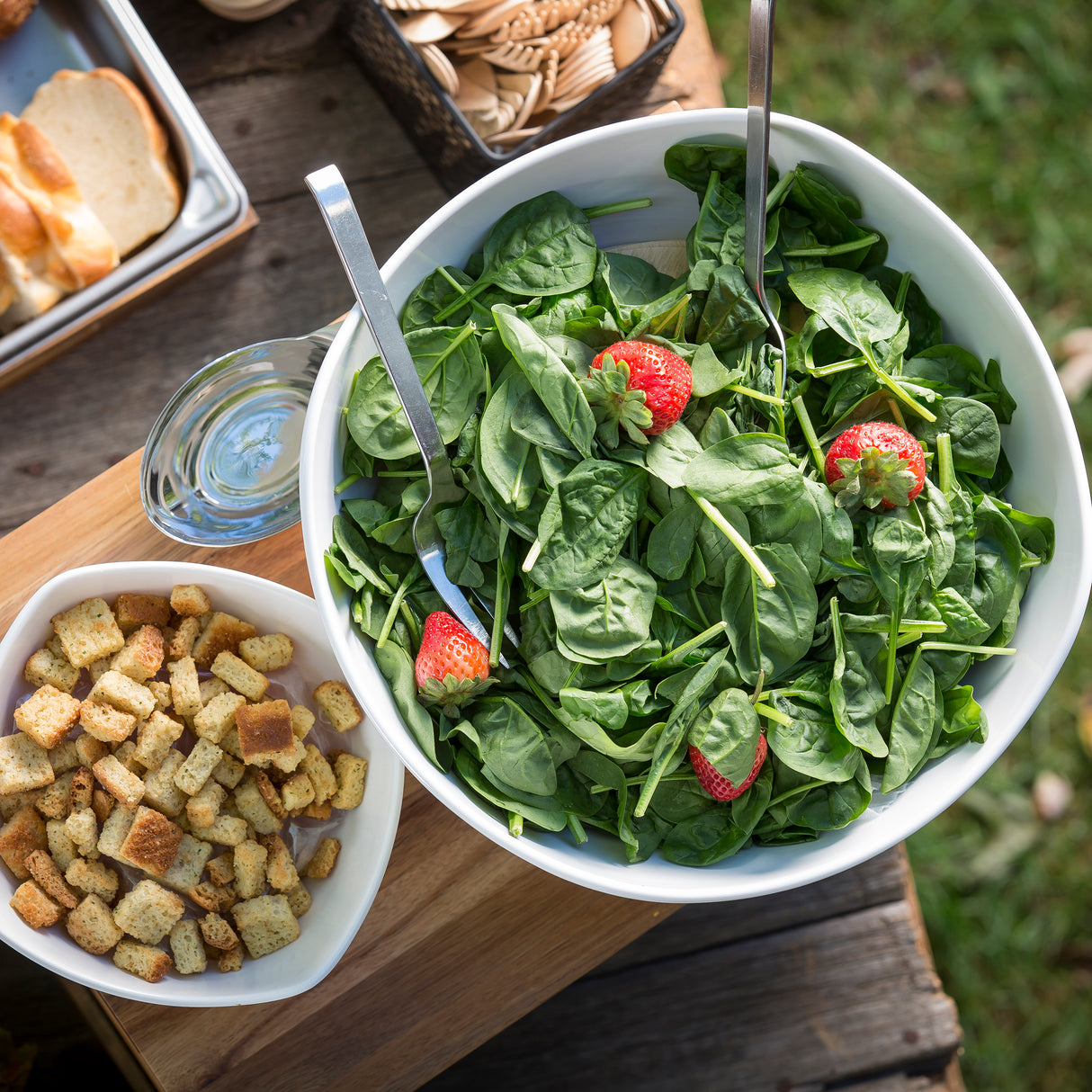 A white melamine bowl from the Frostone Collection Wavy Bowl, filled with fresh spinach and strawberries, sits with serving utensils. Nearby are croutons, sliced baguette, and a glass of water on a rustic outdoor table—all dishwasher safe.