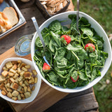 A white melamine bowl from the Frostone Collection Wavy Bowl, filled with fresh spinach and strawberries, sits with serving utensils. Nearby are croutons, sliced baguette, and a glass of water on a rustic outdoor table—all dishwasher safe.