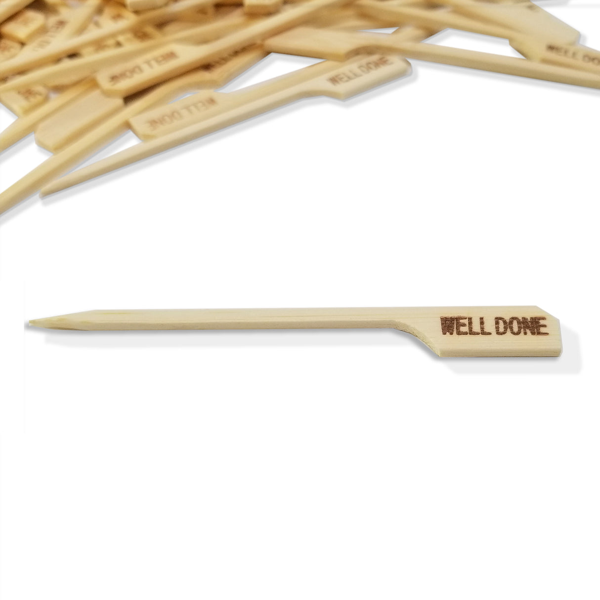 A bamboo meat marker pick lies horizontally on a white surface, engraved with the words “WELL DONE.” More identical picks, some also marked “WELL DONE,” are scattered in the blurred background at the top of the image.