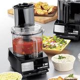 A batch bowl food processor filled with chopped tomatoes sits on a kitchen counter. Nearby are dishes with salad, bread, dip, and a second commercial food processor, suggesting meal prep or serving with fresh ingredients and appetizers.