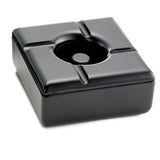 A square black ceramic ashtray with rounded edges and four grooves for holding cigarettes, photographed on a white background. This windproof ashtray features a glossy surface that reflects light, and its recessed center is designed to catch ashes.