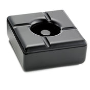 A square black ceramic ashtray with rounded edges and four grooves for holding cigarettes, photographed on a white background. This windproof ashtray features a glossy surface that reflects light, and its recessed center is designed to catch ashes.