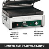 A stainless steel Panini grill with ribbed cast iron plates, timer knob, power and ready indicator lights, and an adjustable thermostat up to 570°F. A label notes its limited one-year warranty. The grill lid is open, revealing the interior plates.