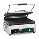 A stainless steel Waring Panini grill with black ribbed cast iron plates, open lid, and green control panel. The panel features an adjustable thermostat, power switch, and indicator lights. The handle is black and silver. The grill is clean and unused.