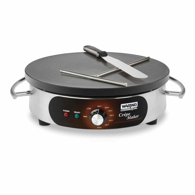 A stainless steel Waring Commercial Crepe Maker with a large round cast iron cook surface, two metal handles, a batter spreader, and spatula. The front panel features an adjustable thermostat, indicator lights, and labeled controls.