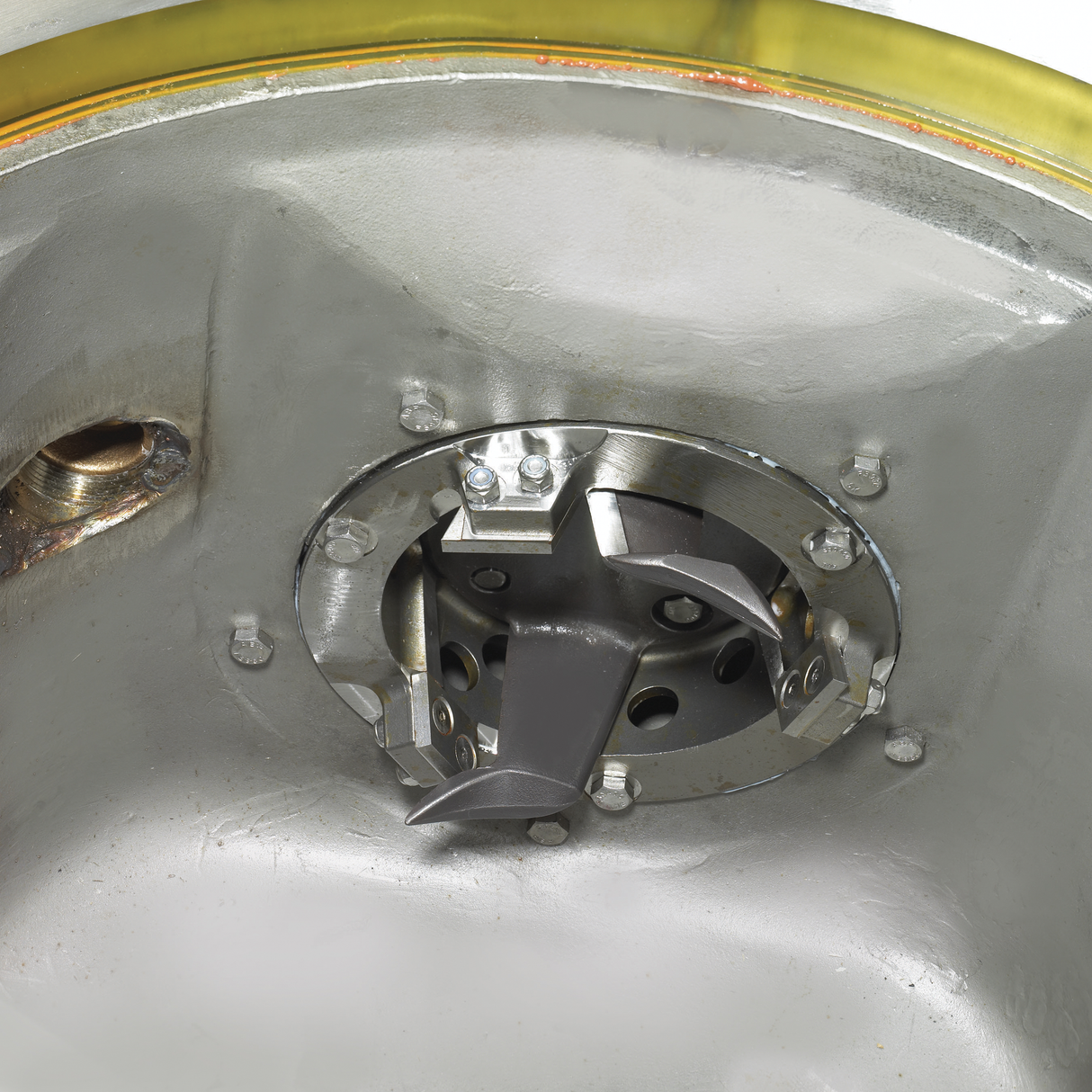 Close-up of a metallic circular mechanism, possibly part of a WastePro self-contained pulper, with visible bolts, curved blades, and perforated inner sections, mounted inside a smooth metallic surface featuring a yellowish translucent edge at the top.