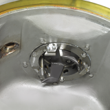 Close-up of a metallic circular mechanism, possibly part of a WastePro self-contained pulper, with visible bolts, curved blades, and perforated inner sections, mounted inside a smooth metallic surface featuring a yellowish translucent edge at the top.