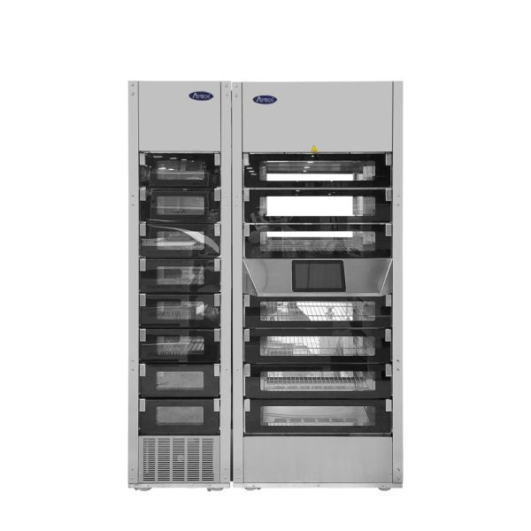 Two tall, stainless steel vending machines stand side by side. One is a Heated Food Locker with a digital touchscreen in the center, while both have transparent-front compartments. The machines boast a modern, industrial appearance.