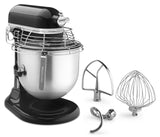 A black and silver KitchenAid Commercial Stand Mixer with an 8 quart bowl and wire safety guard is shown. Three metal attachments—a flat beater, dough hook, and wire whisk—are placed beside it on a plain white background.