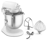 A white KitchenAid Commercial Stand Mixer with a stainless steel 8 quart bowl and protective wire guard. Three metal attachments—a flat beater, dough hook, and wire whisk—are displayed in front on a white surface.