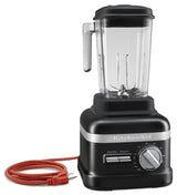 A black KitchenAid Commercial Bar Blender with a clear plastic pitcher and handle, patented Talon asymmetrical blade, silver control dial, “Start/Stop” and “Pulse” buttons, and a red power cord coiled beside the base. Shown on a white background.