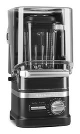 A black and silver KitchenAid Commercial Blender with a 60 oz blending jar features a clear noise-reducing enclosure. Its control panel includes start/stop, pulse/variable speed options, and a large dial marked from low to high.
