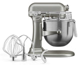 A silver KitchenAid Commercial Stand Mixer with an 8 quart bowl and stainless steel mixing bowl attached. In front are three metal attachments: a wire whisk, flat beater, and dough hook. The mixer has a safety guard and lever on the side.