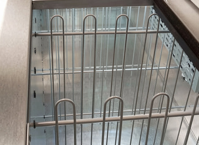 A close-up view of the interior of an empty Arctic Air freezer or refrigerated display case, showing shiny metallic surfaces and white wire racks with a vertical can divider. The image is taken from above, looking down into the clean AUB50R/65R unit.