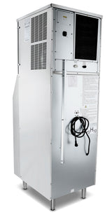 A large, silver, rectangular hotel ice machine with vent panels, a control panel, and a black cord hanging on the front stands on four metal legs. Labels, warning signs, and various screws and panels mark this full cube ice dispensers surface.