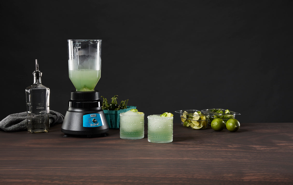 A Bar Blender with a 48 oz. capacity holds a light green drink on a wooden surface next to two filled glasses with lime wedges, a bottle with a spout, mint leaves in a blue container, and sliced limes on a tray. The background is dark.