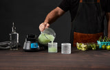 A person in a black apron pours a green smoothie from a blender with a stainless steel blade into two textured glasses on a wooden table. Nearby are the Bar Blender base, bottle, towel, limes, sliced limes, and green chili peppers against a dark background.