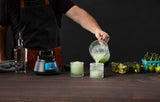 A person in a black shirt and apron pours a green blended drink from a pitcher into two textured glasses on a wooden table, surrounded by a Bar Blender with stainless steel blade, limes, sliced peppers, and containers of garnish. Background is dark.