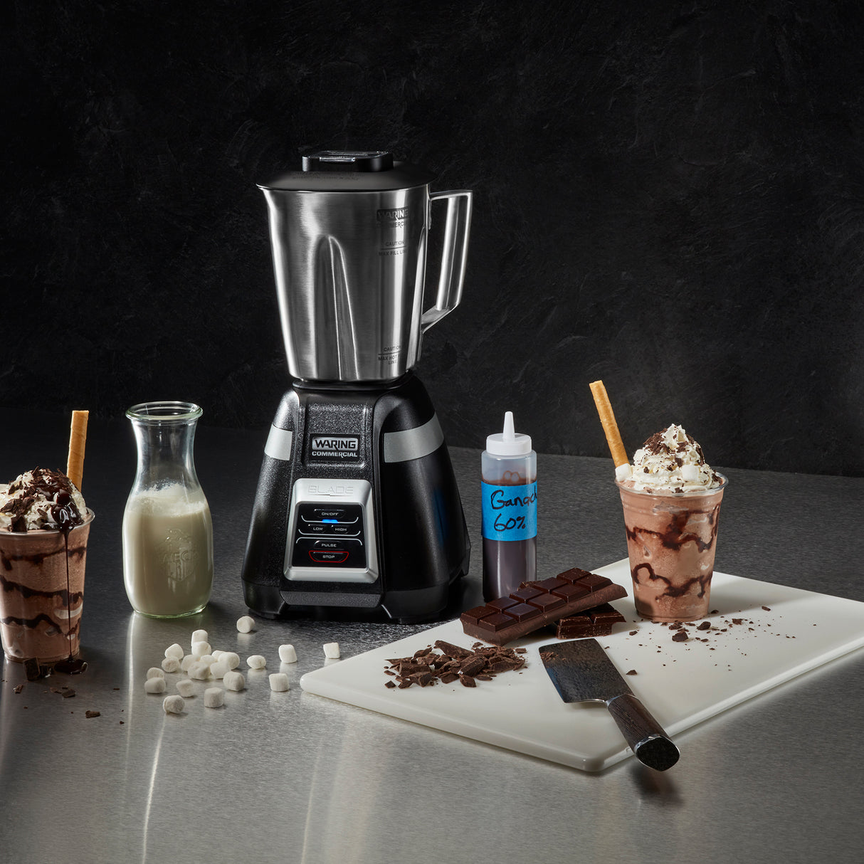 A stainless steel bar blender sits on a counter with a bottle of milk, chocolate bars, marshmallows, a squeeze bottle labeled “GARNISH,” a cutting board with chopped chocolate and a knife, and two chocolate milkshakes topped with whipped cream and wafers.