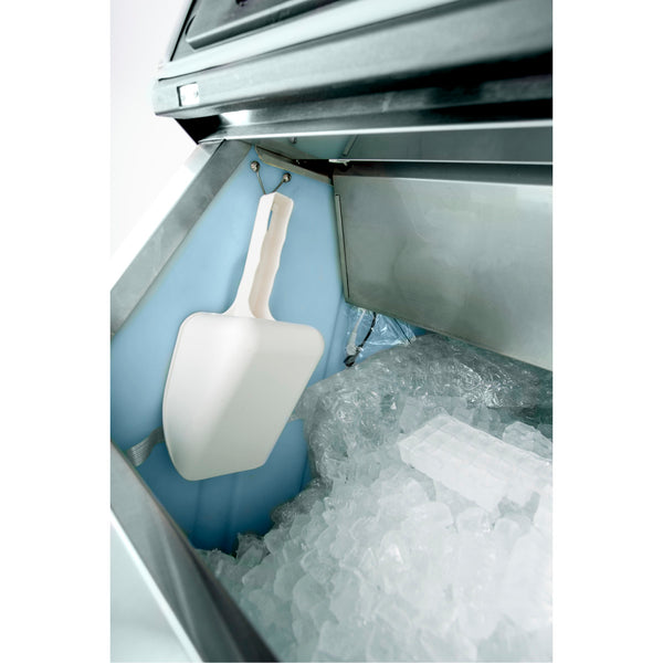A white plastic ice scoop hangs on a hook inside an industrial stainless steel ice maker filled with clear ice cubes. The blue metallic interior and an ice storage bin are visible with the machine’s lid open, revealing its contents.