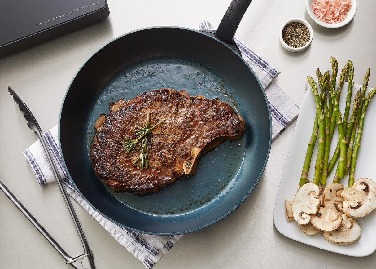 A seared steak with rosemary sits in a SwissSteel Fry Pan on a striped cloth. Beside it are tongs, bowls of salt and pepper, and a white plate with asparagus and sliced mushrooms, all on a light countertop.