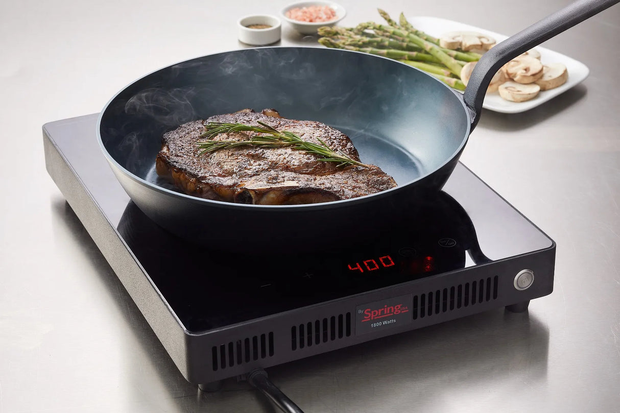 A seared steak with a sprig of rosemary cooks in a black carbon steel fry pan on an induction cooktop set to 400°F. Steam rises from the pan, while plates with asparagus, mushrooms, and seasoning sit on a stainless steel countertop.