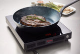 A thick steak, topped with rosemary, sizzles in a Blackline SwissSteel Fry Pan on a black induction cooktop displaying 400. In the background, fresh asparagus, sliced mushrooms, and coarse pink salt await. Steam rises from the steak.