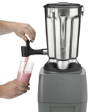 A person’s hands pour a pink smoothie from a spout attached to a heavy-duty Food Blender with a stainless steel container into a clear glass. The industrial-sized blender, on a gray base, is the focus as the smoothie streams smoothly.