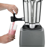 A person uses a lever spout to pour a pink smoothie from a heavy-duty blender with a stainless steel container into a clear glass. One hand operates the lever while the other holds the glass; the blender sits on a gray base against a plain white background.