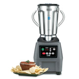A commercial-grade Waring Food Blender with a heavy-duty digital control panel and stainless steel container sits on a white surface. In front, a plate of tortilla chips and a bowl of salsa garnished with herbs complete the scene.
