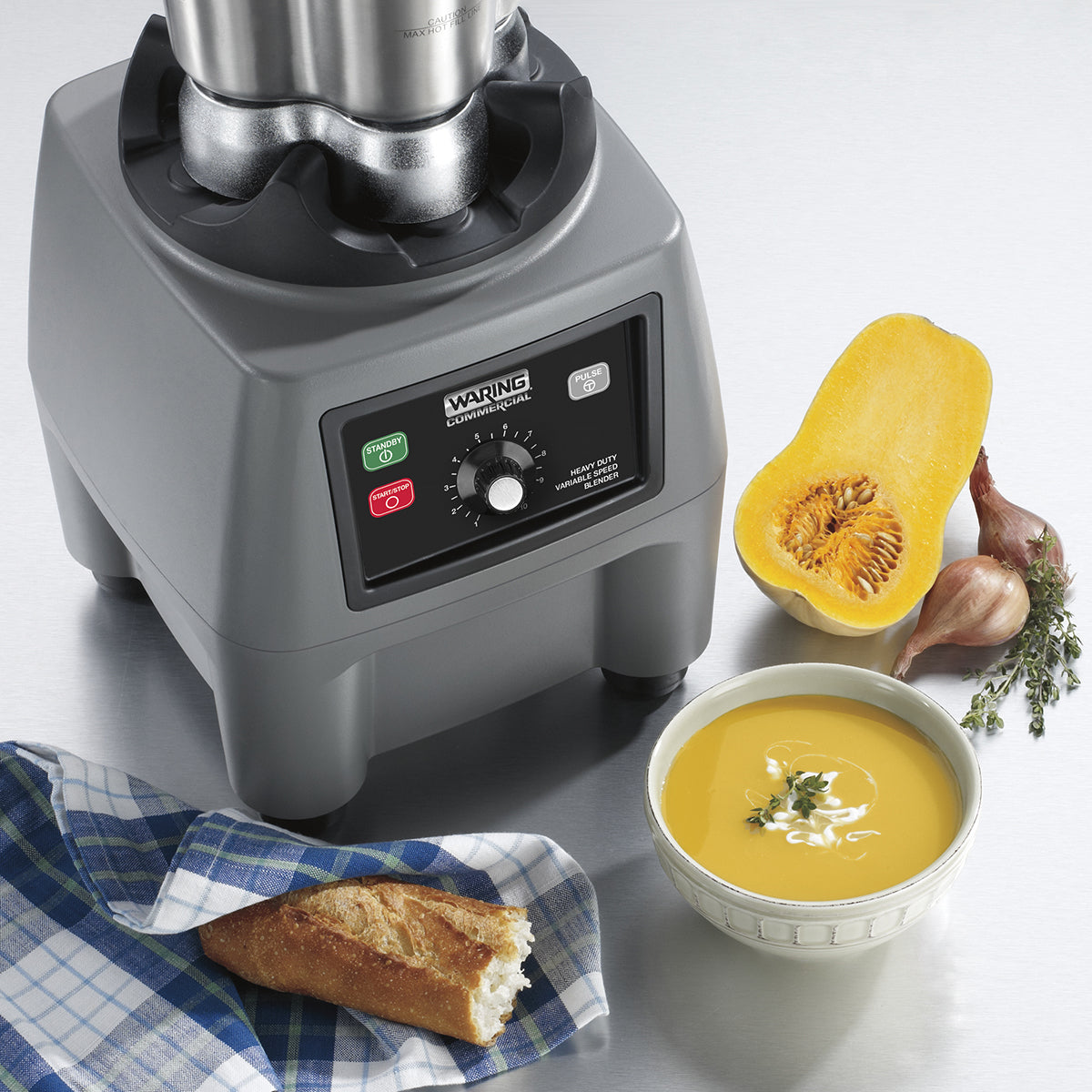 A gray commercial blender base sits on a metal counter beside a halved butternut squash, two shallots, fresh herbs, a bowl of creamy soup with garnish, and a piece of baguette on a blue-patterned cloth.