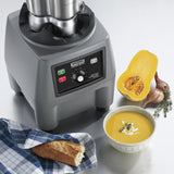 A gray commercial blender base sits on a metal counter beside a halved butternut squash, two shallots, fresh herbs, a bowl of creamy soup with garnish, and a piece of baguette on a blue-patterned cloth.