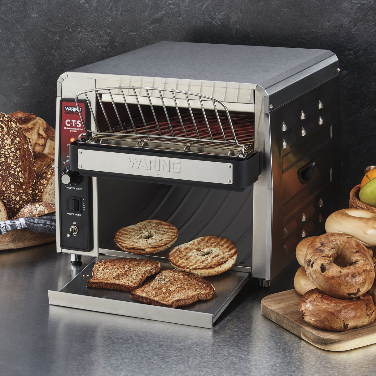 A stainless steel Waring conveyor toaster oven with a wire rack and open front is toasting bread slices and bagels. Around the toaster, various breads, bagels, and fruit are arranged on wooden boards against a dark background.