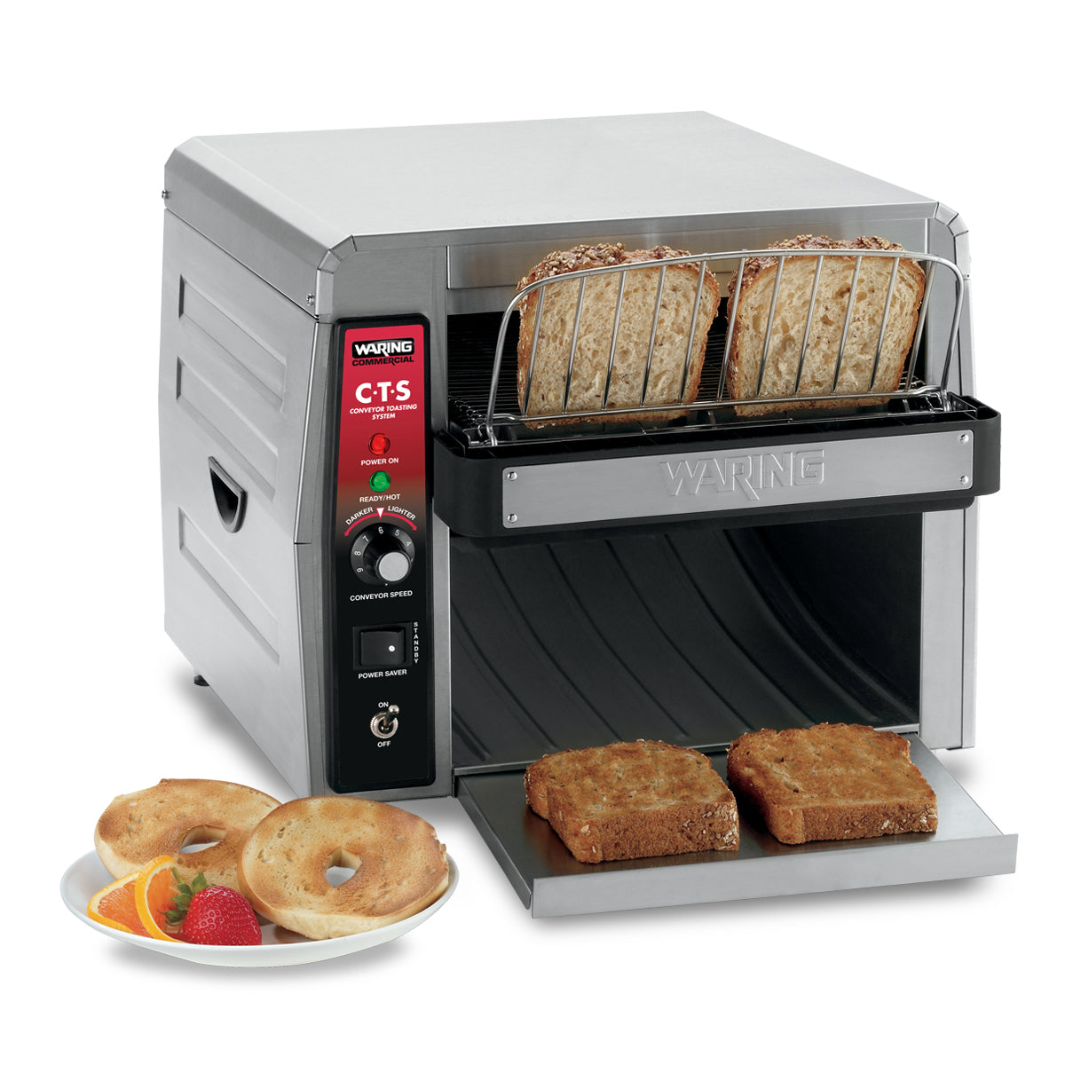 A stainless steel Waring CTS1000AL conveyor toaster with control dials toasts two slices of bread. Two toasted slices emerge on a tray. Beside it, a plate holds two bagel halves and mixed fruit, including a strawberry and melon slices.
