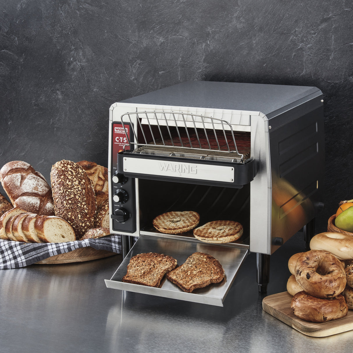 A commercial conveyor toaster on a metal countertop toasts bread slices. Toasted bread slides onto a tray, while various breads—sliced loaves, rolls, buns, and bagels—are arranged beside the stainless steel toaster. The background is a dark textured wall.