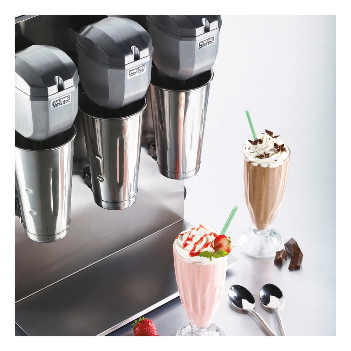 A triple spindle milkshake machine with three metal blending cups sits on the countertop. In front are two milkshakes—one pink with whipped cream and strawberries, one brown with whipped cream and chocolate shavings—plus spoons and chocolate pieces.