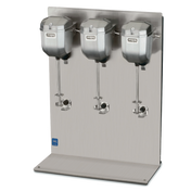 A stainless steel triple spindle drink mixer with three large mixing heads, each with a spindle and beater, mounted on a vertical metal backing plate. The countertop base is flat and polished. The Waring Commercial brand name is visible.