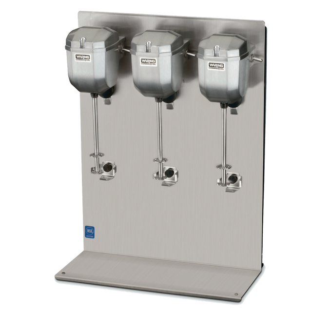 A stainless steel triple spindle drink mixer with three large mixing heads, each with a spindle and beater, mounted on a vertical metal backing plate. The countertop base is flat and polished. The Waring Commercial brand name is visible.