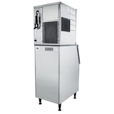 A commercial ice machine with a boxy, stainless steel body stands on four legs. The top section has vents, an open maintenance panel with coiled tubing, and instruction labels, while the lower section features a commercial ice storage door and handle.