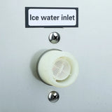A close-up of a white plastic Ice water inlet on a light gray surface, commonly found on a commercial ice maker. The inlet features a mesh filter inside, with two metallic screws above and below it securing it in place.